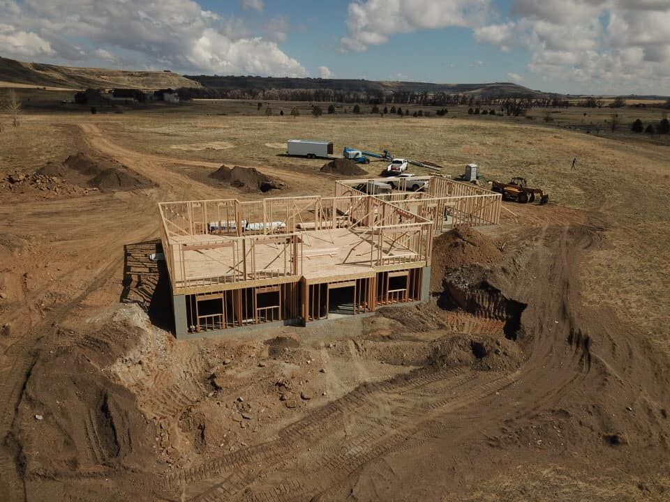 House under construction in a field, with wooden frame, dirt, and construction equipment.