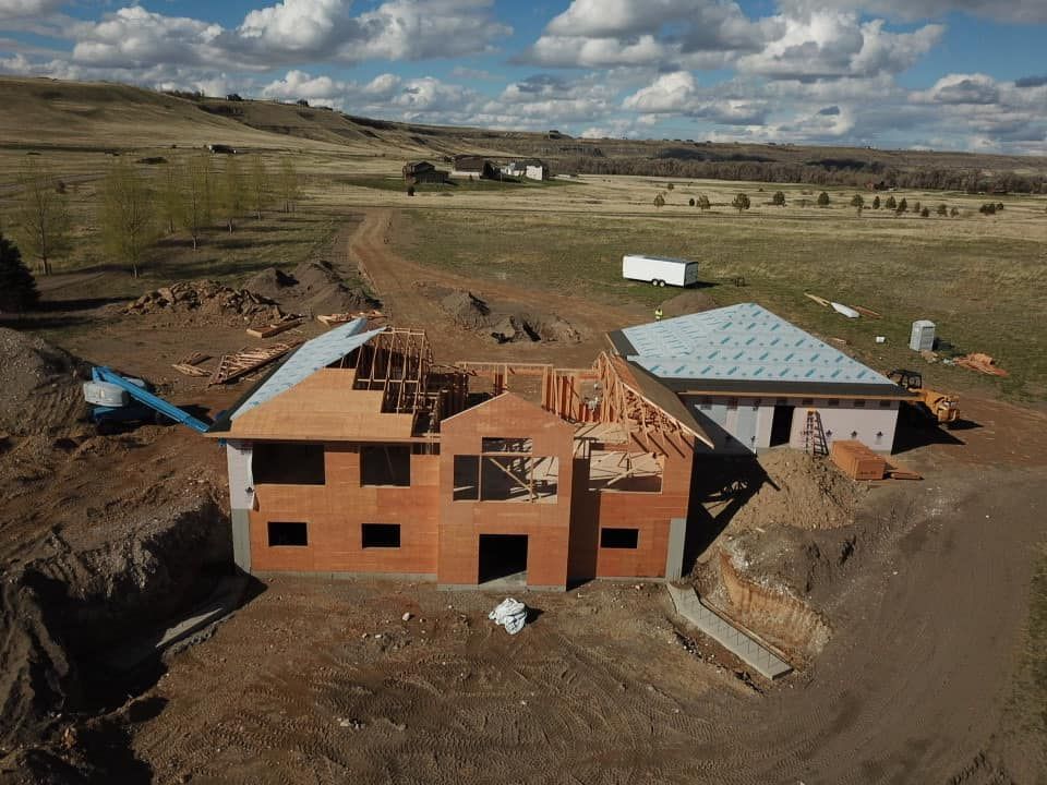Construction site: framing a house with exposed wooden beams, brown siding, in a rural, hilly landscape under a blue sky.