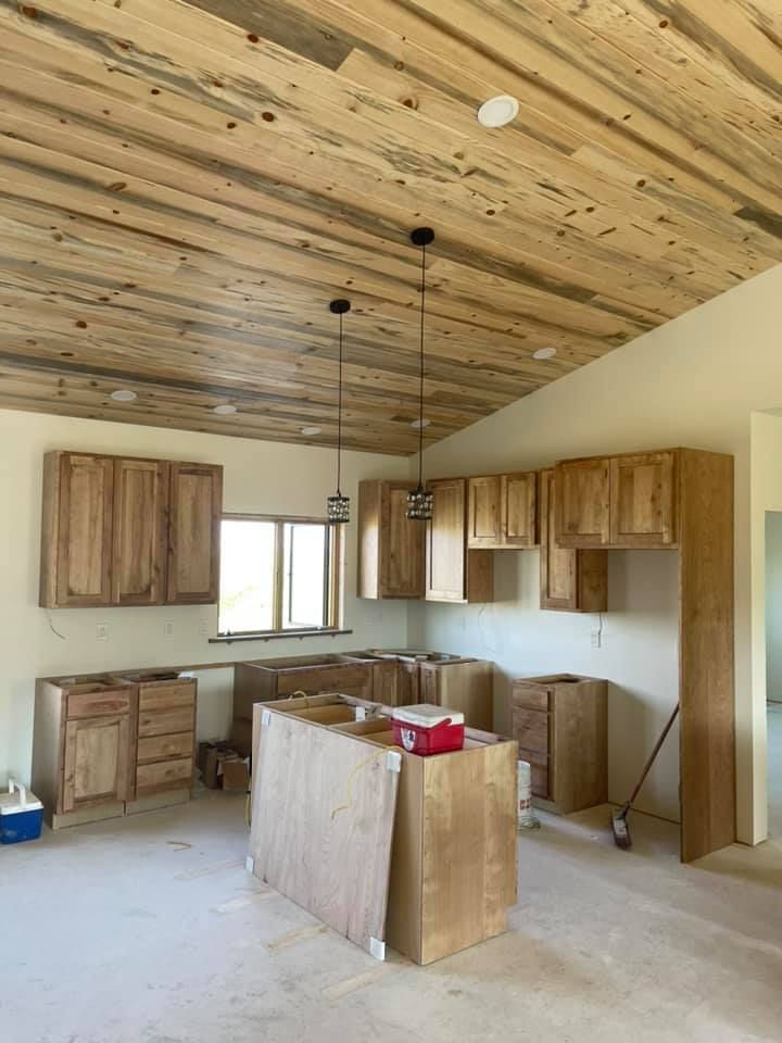 Unfinished kitchen with wood cabinets, island, and ceiling. Two pendant lights hang. Window visible.