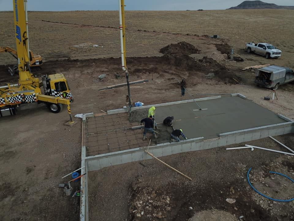Construction workers pouring concrete foundation for a building in an open field. A yellow concrete pump truck is visible.