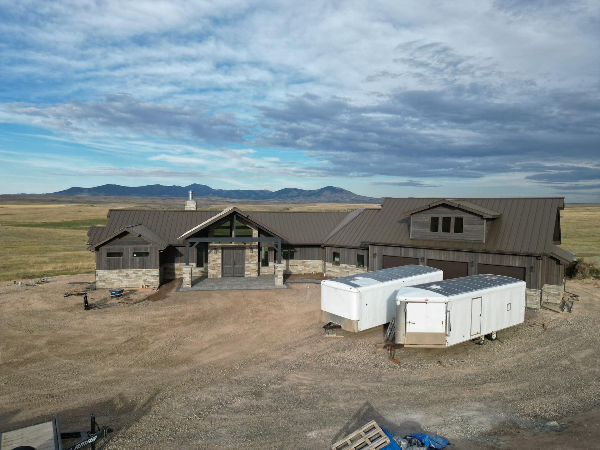 A large, rustic house under construction with white trailers in front, set in a vast, open landscape.