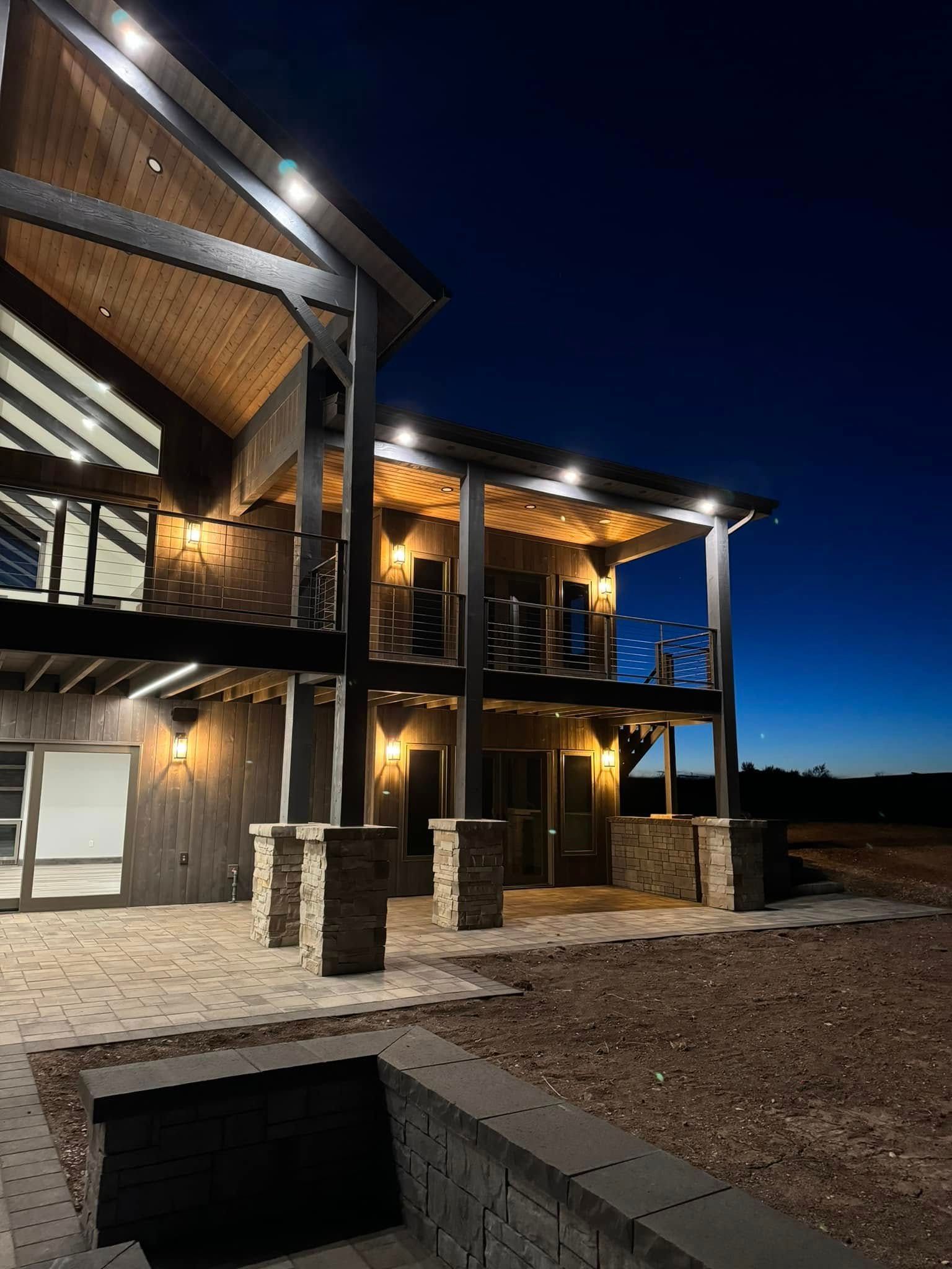 Two-story house exterior lit at dusk. Wooden beams, stone columns, and outdoor lighting against a dark blue sky.