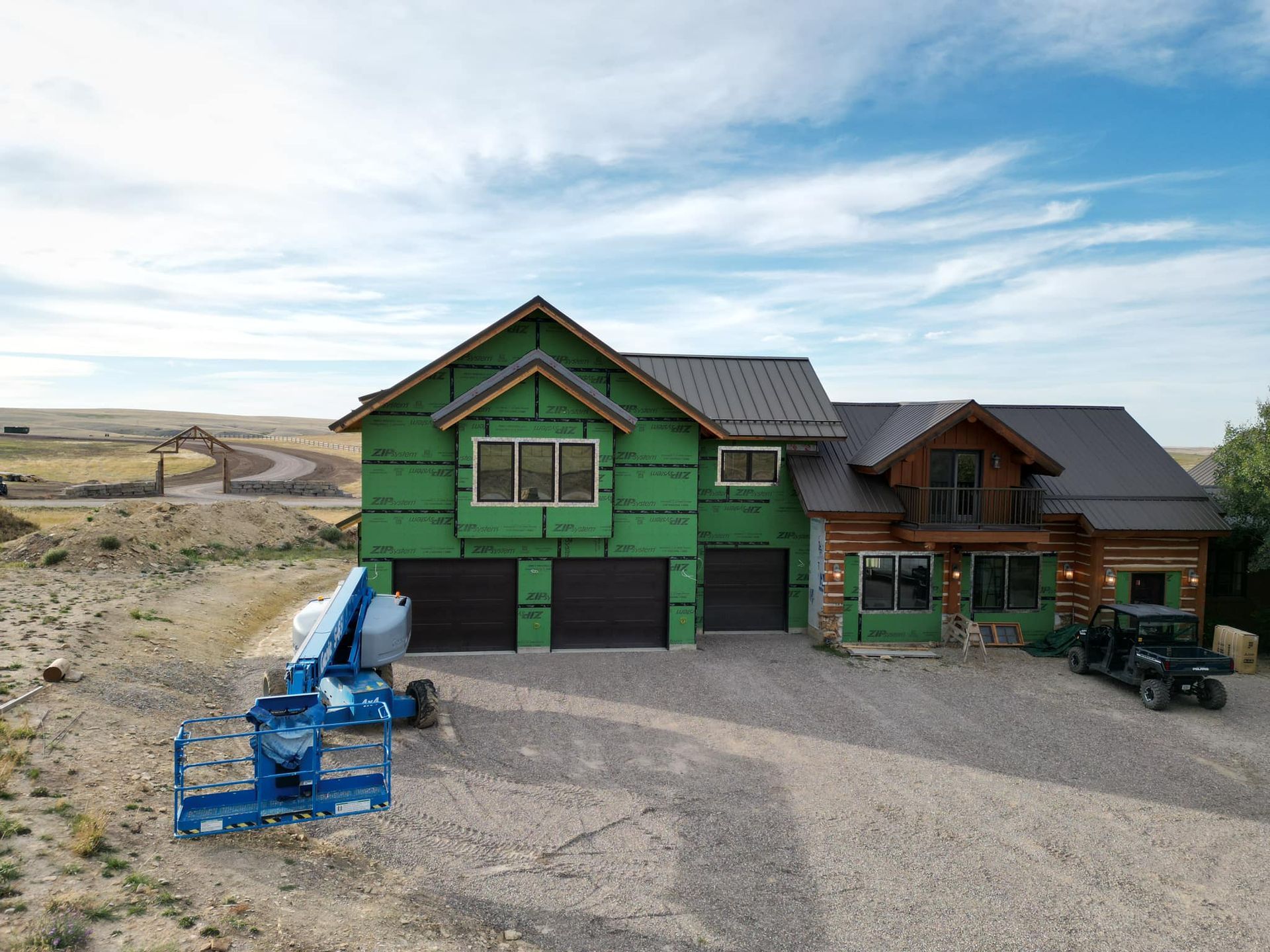 A house under construction; green exterior wrap, brown roof, and gravel driveway. A blue lift is in front.