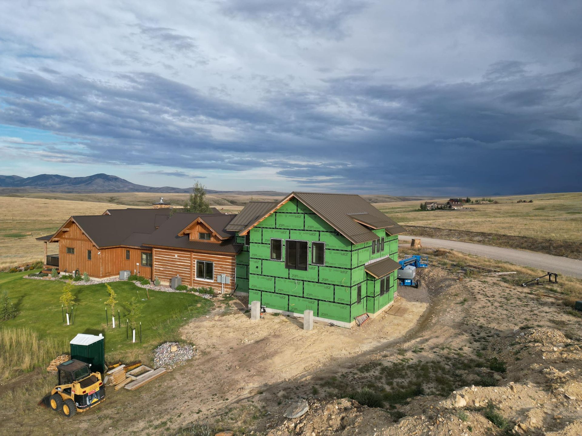 House under construction, with brown roof on left section and green wrap on right, set in a rural landscape.