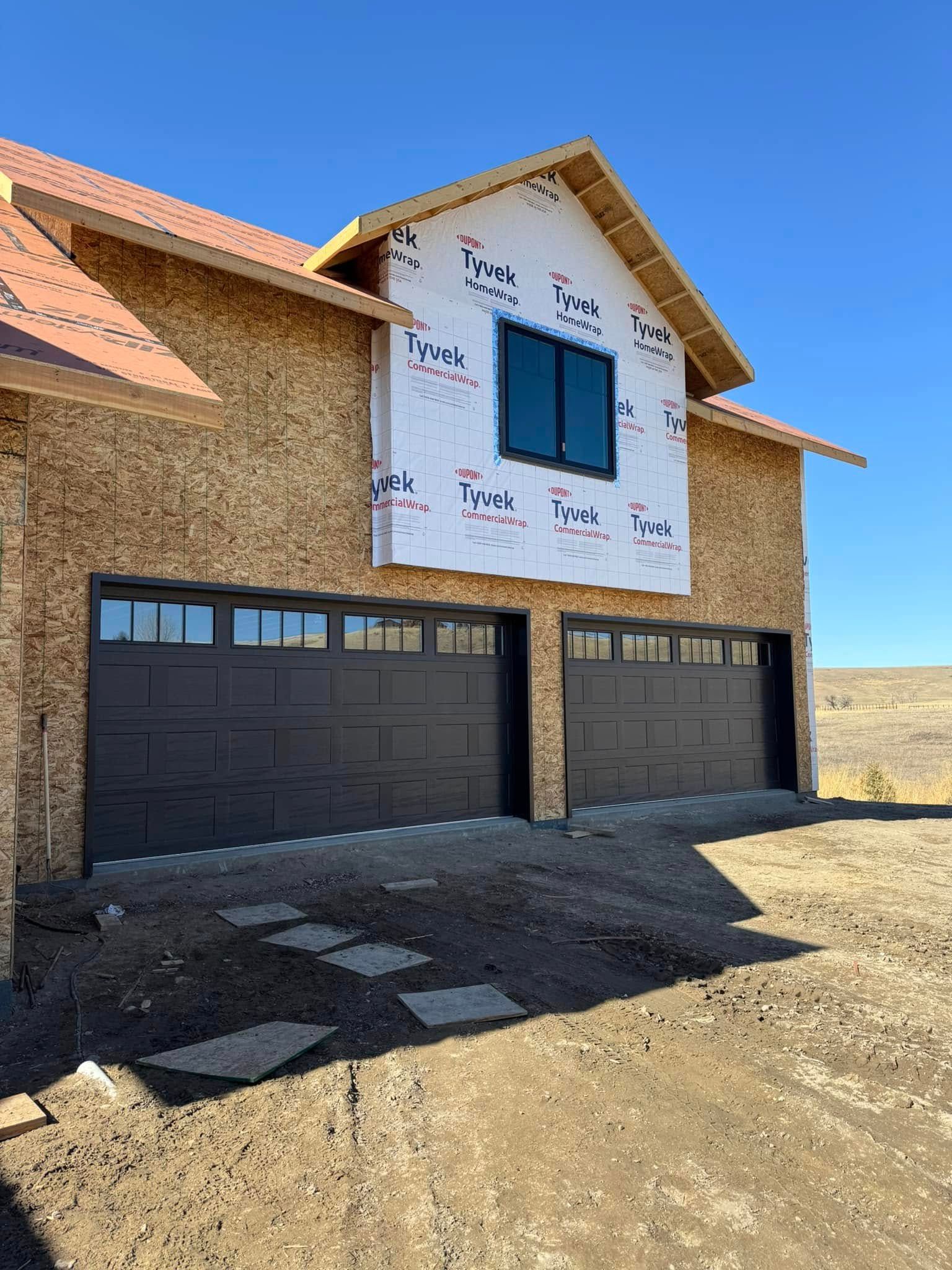 Garage doors on a new house under construction. Dark gray doors, unfinished roof, and blue sky.
