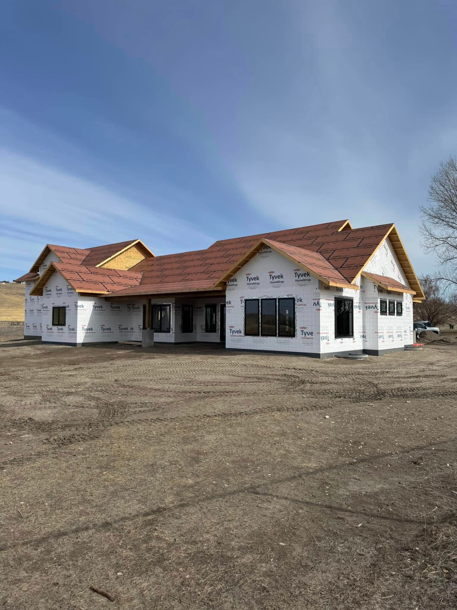 New home construction with brown roof and black framed windows, covered in white wrap, on a dirt lot under blue sky.