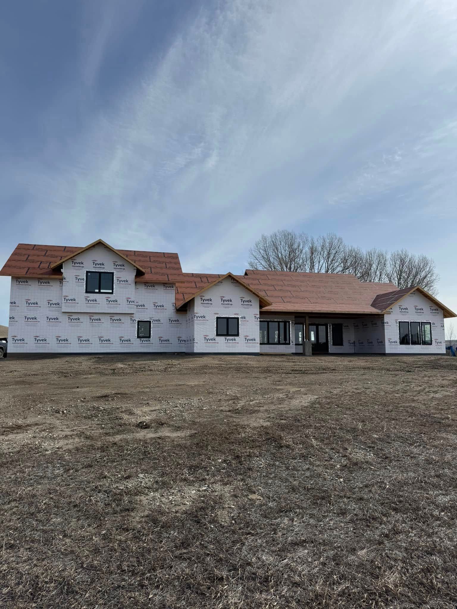 New house under construction with brown roof, windows, and white siding on a dirt lot under a blue sky.