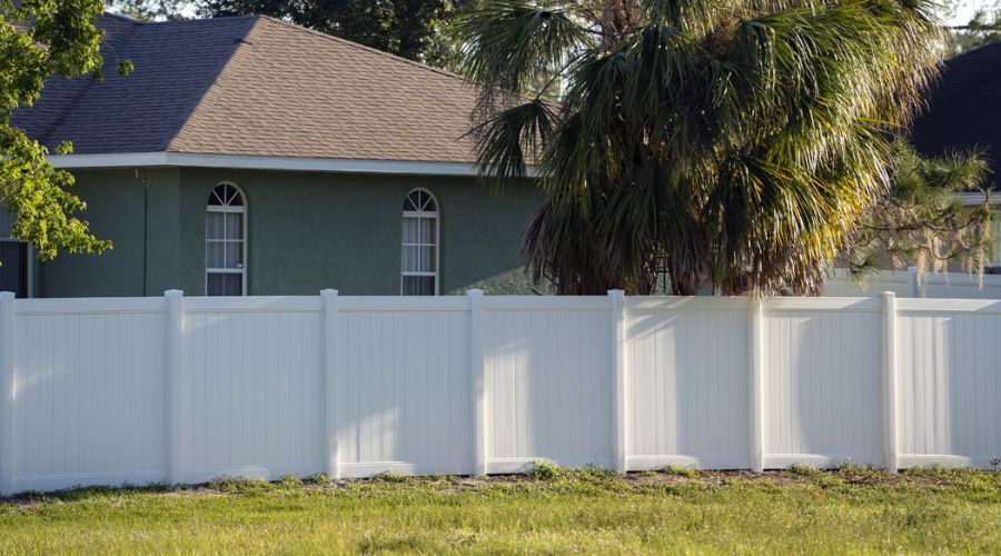 A white vinyl privacy fence stands in a grassy yard in front of a green suburban house with a palm tree.