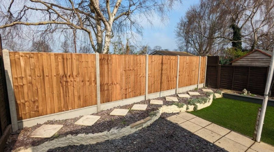 A new wooden fence with concrete posts and gravel boards behind a curved tiered flower bed with paving stones and slate.