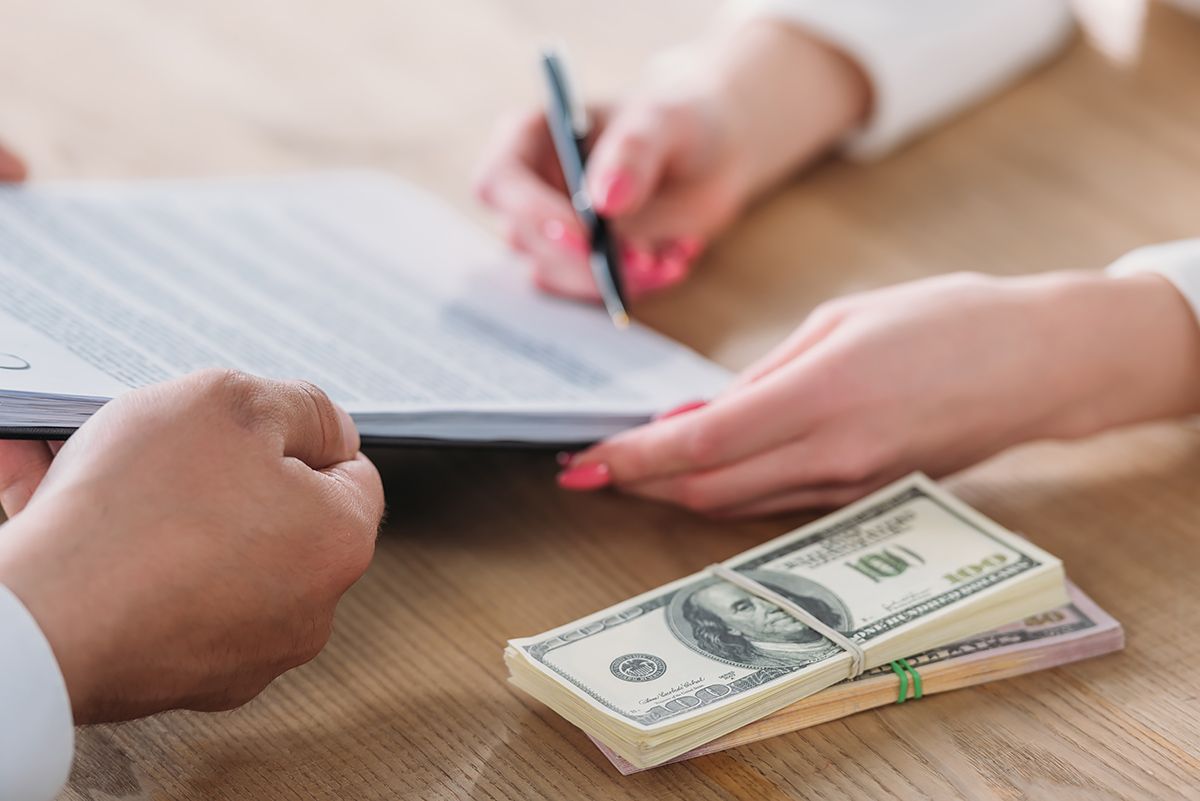 A woman is signing a document next to a stack of money.