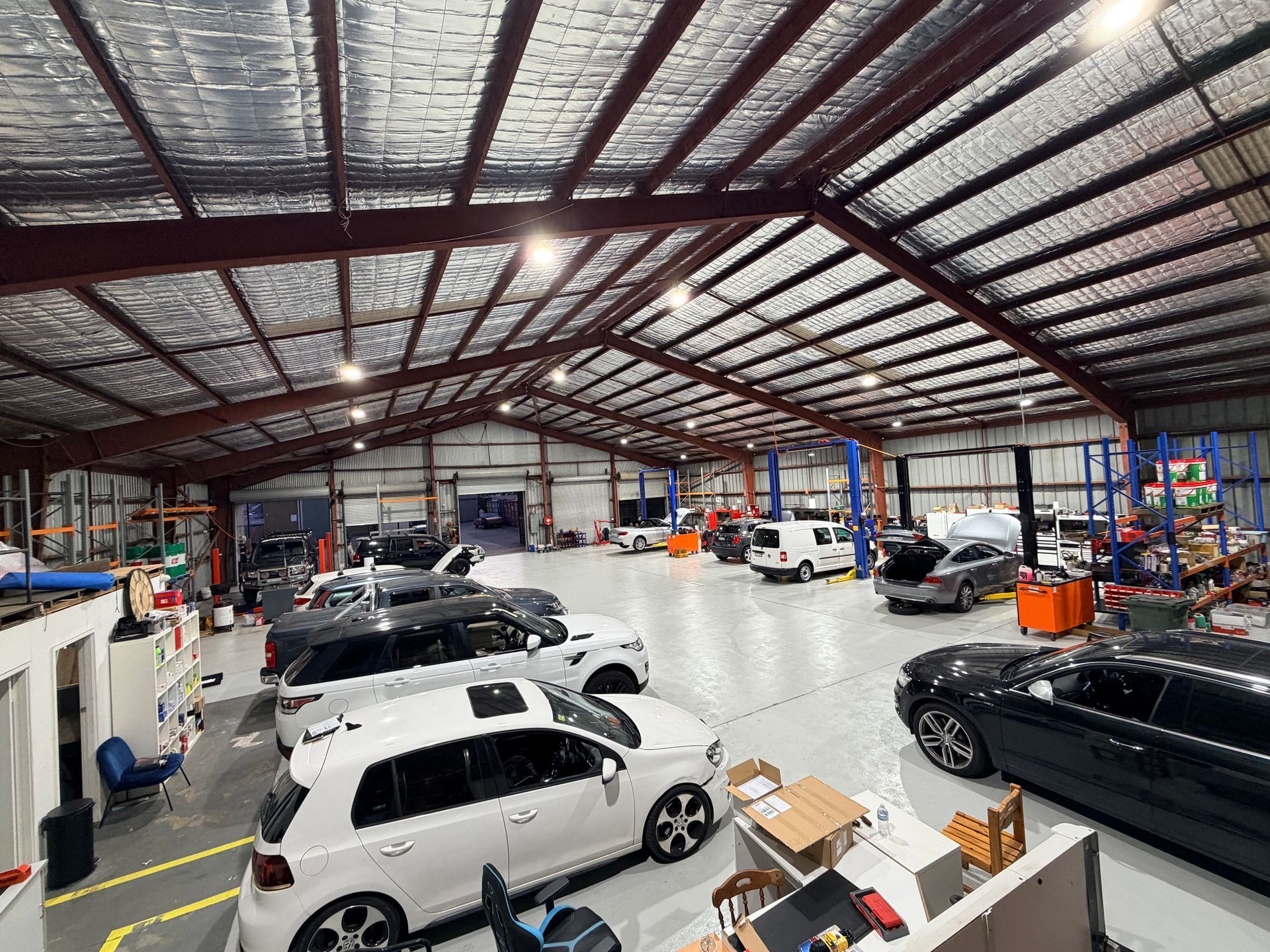 Inside a spacious auto repair shop, several cars are parked, with equipment and shelves lining the walls.