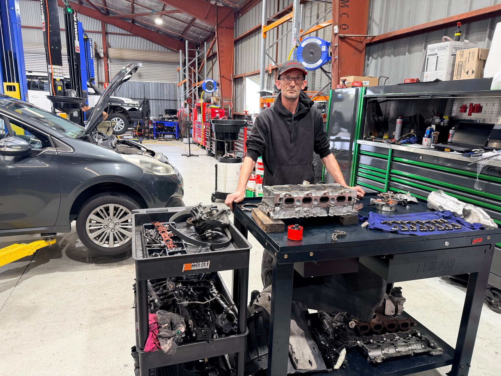 Man in auto shop, smiling, standing near a workbench with car engine parts laid out, car on lift in background.