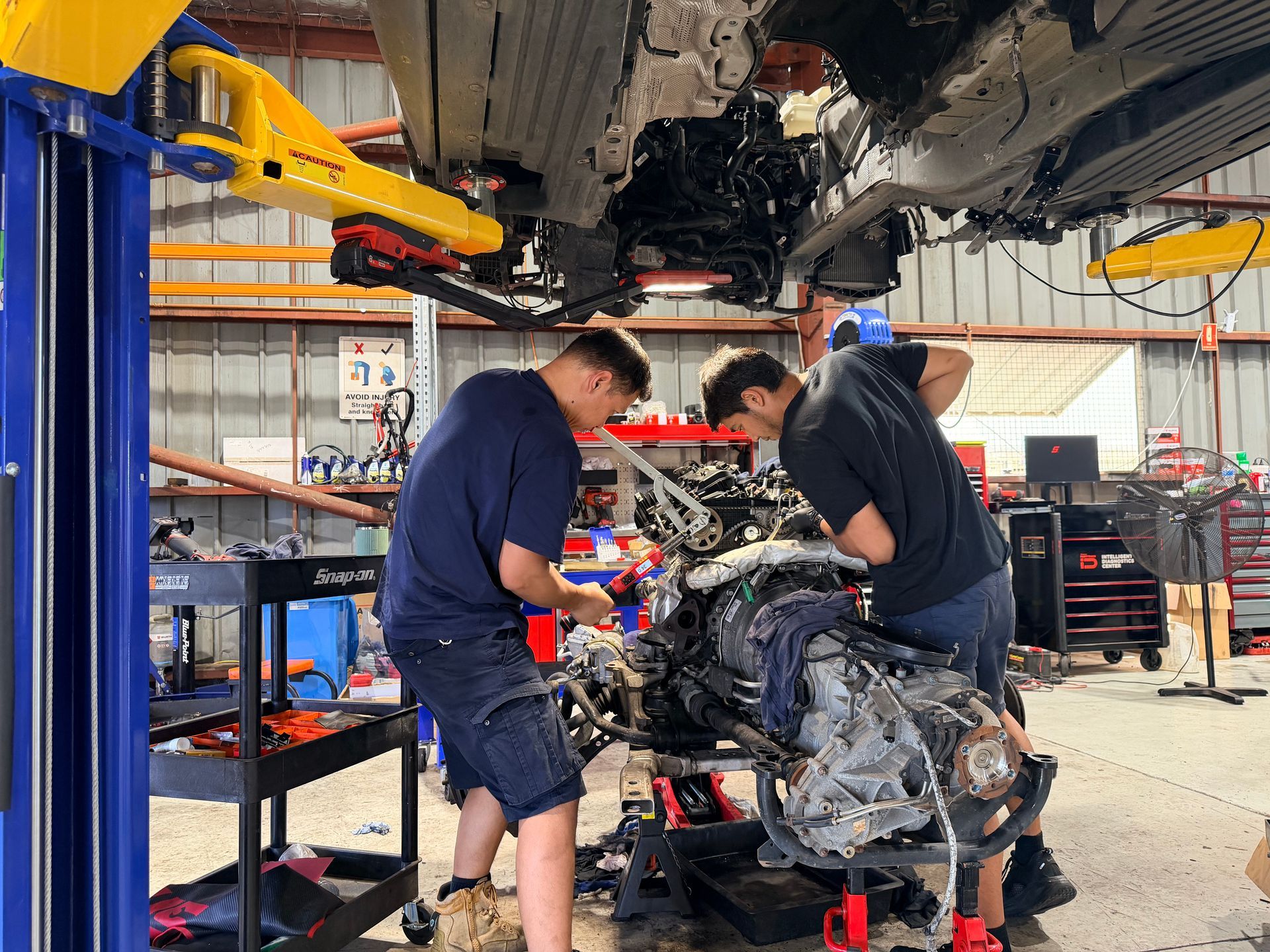 Two mechanics working on a car engine in a garage. They are using tools while the car is raised.