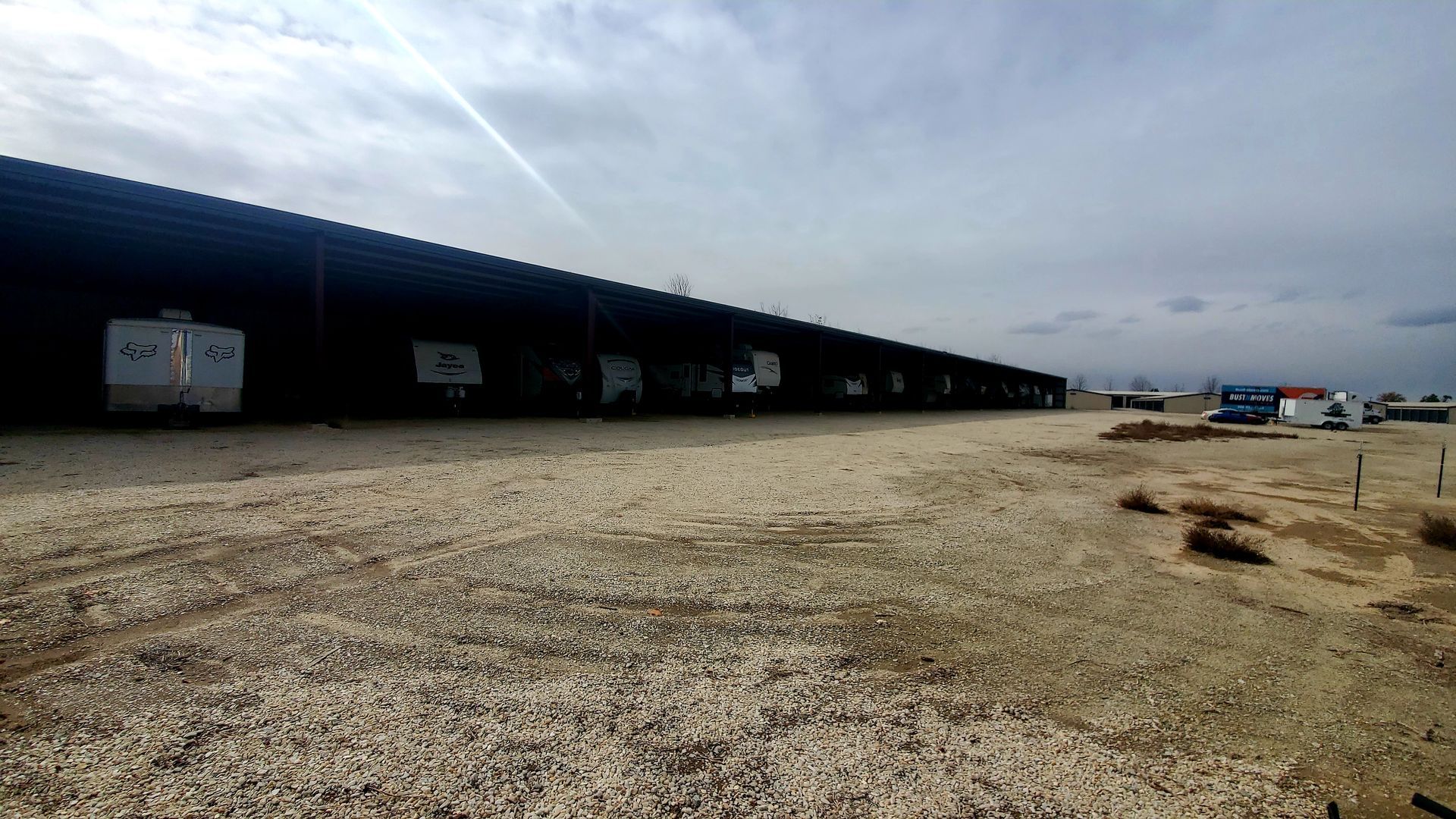 A row of trailers are parked under a roof in a gravel lot.