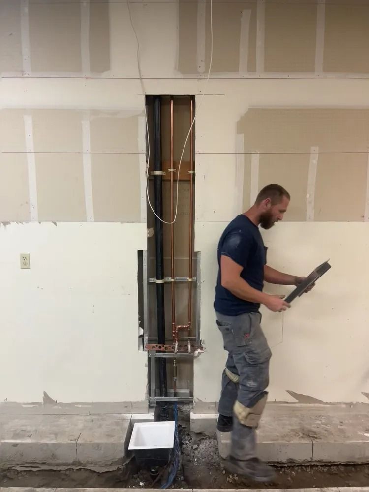 Construction worker inspecting a metal sheet near exposed pipes in a wall cavity, standing next to a trench.
