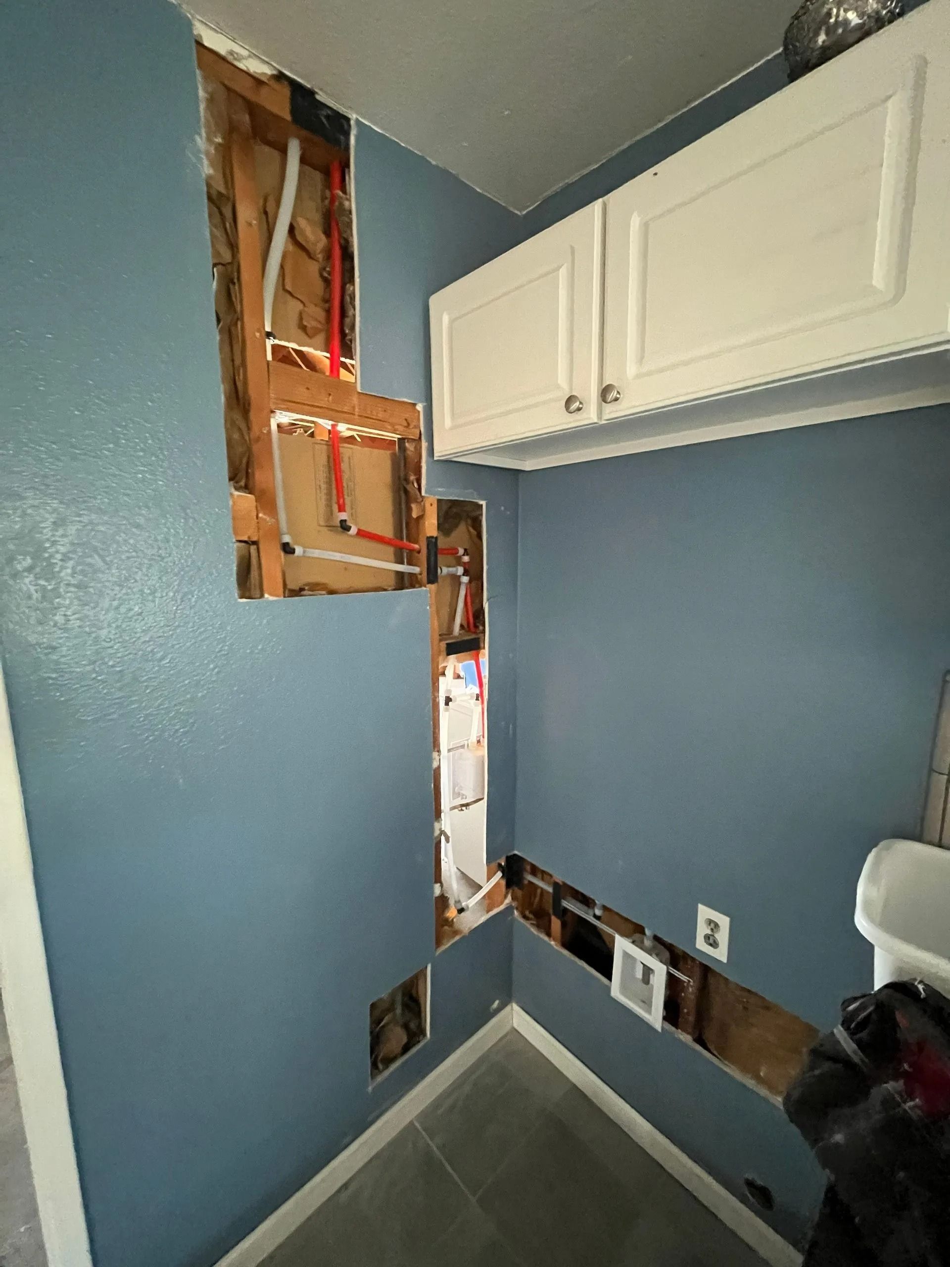 Interior wall with sections removed, exposing plumbing and framing. Blue paint, white cabinet above.