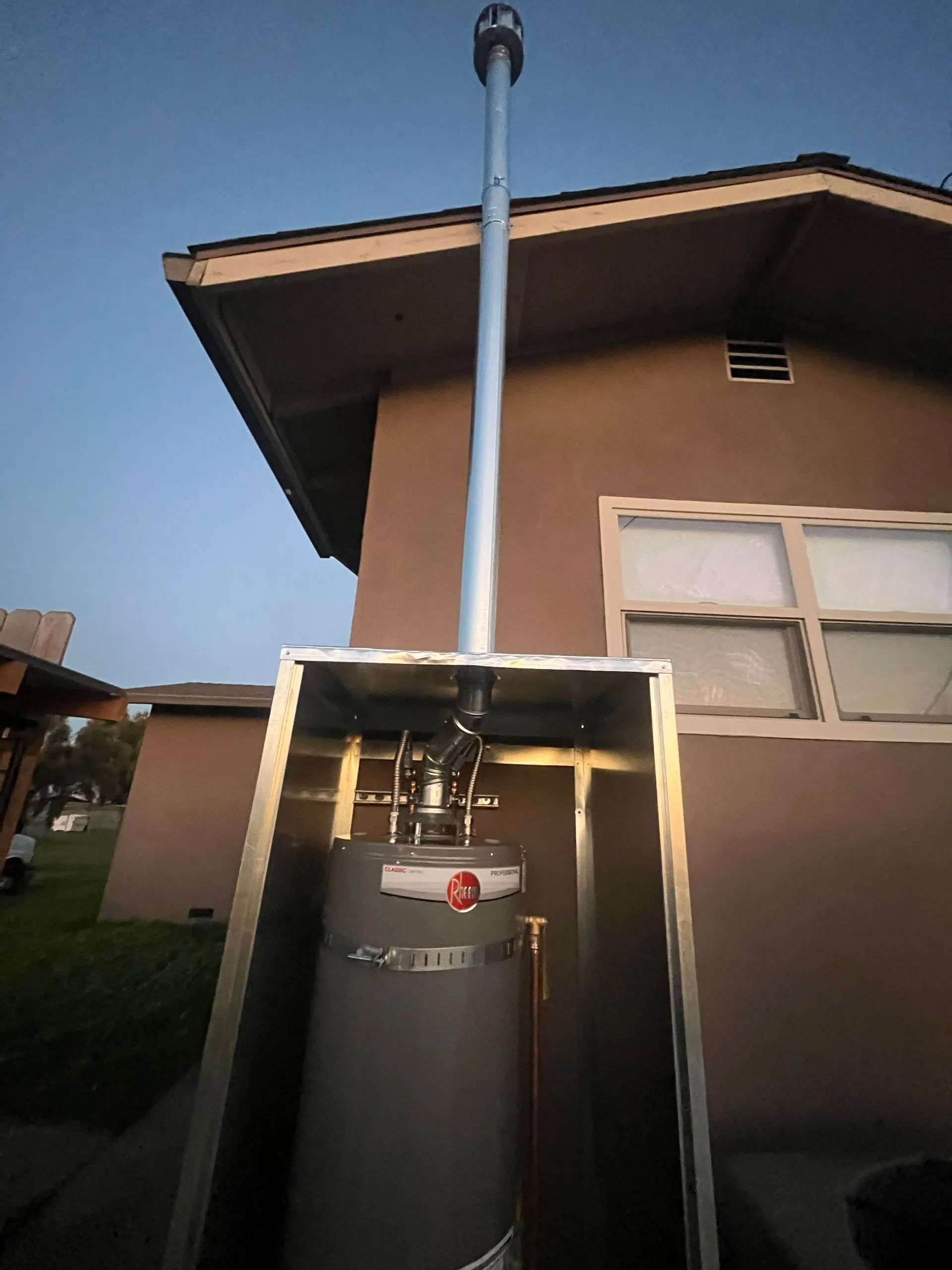 Water heater in metal enclosure with tall flue pipe extending above a house roof.