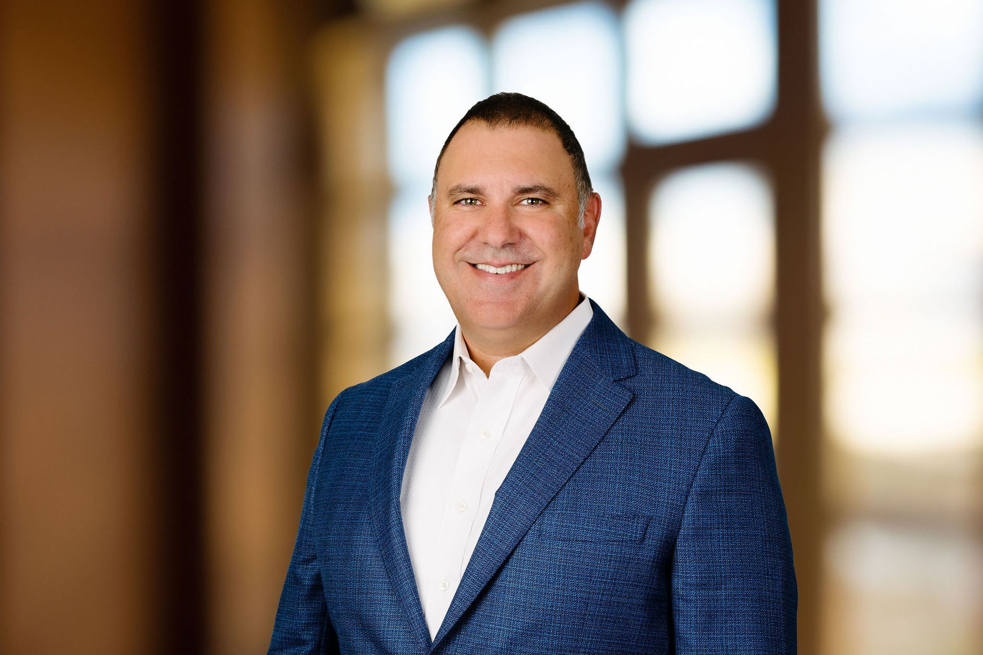 A man in a blue suit and white shirt is smiling in front of a window.