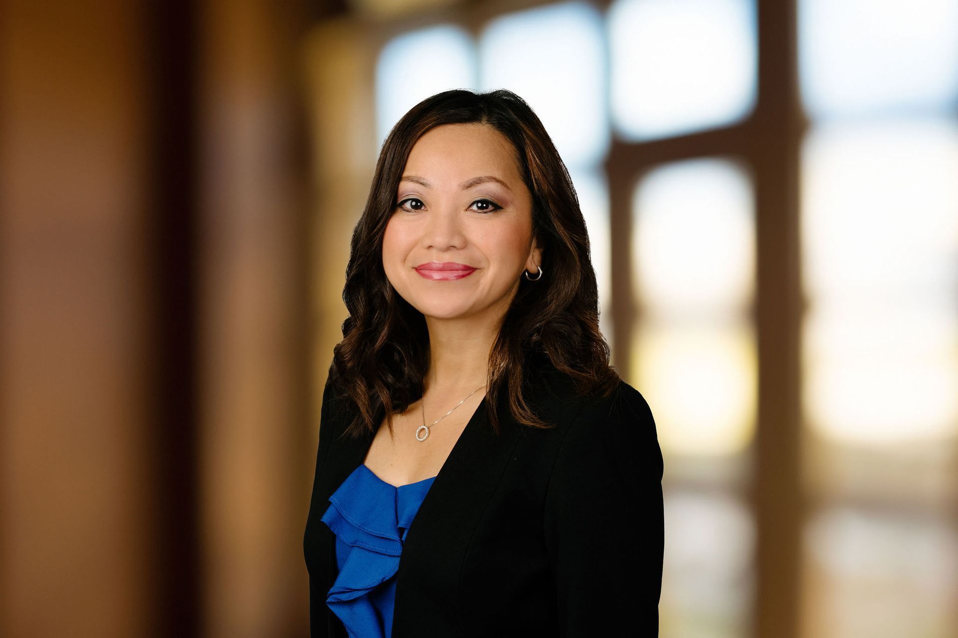 A woman in a blue dress and black jacket is standing in front of a window.
