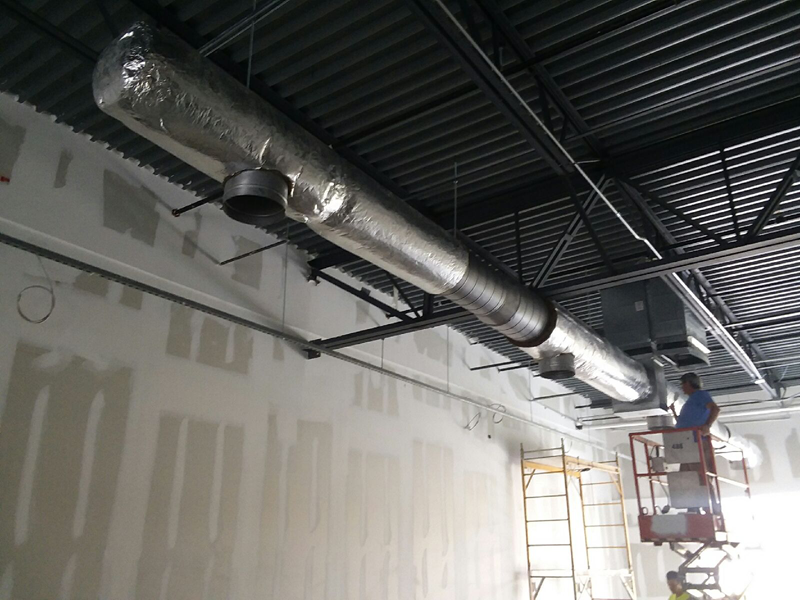 HVAC ductwork installation in a new construction setting. A worker on a lift installs ducting near drywall and black ceiling.