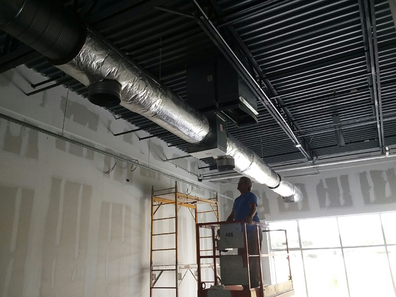 A worker on a lift installs HVAC ductwork in a room with unfinished walls and ceiling.