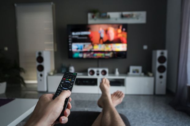 A person is sitting on a couch holding a remote control in front of a television.