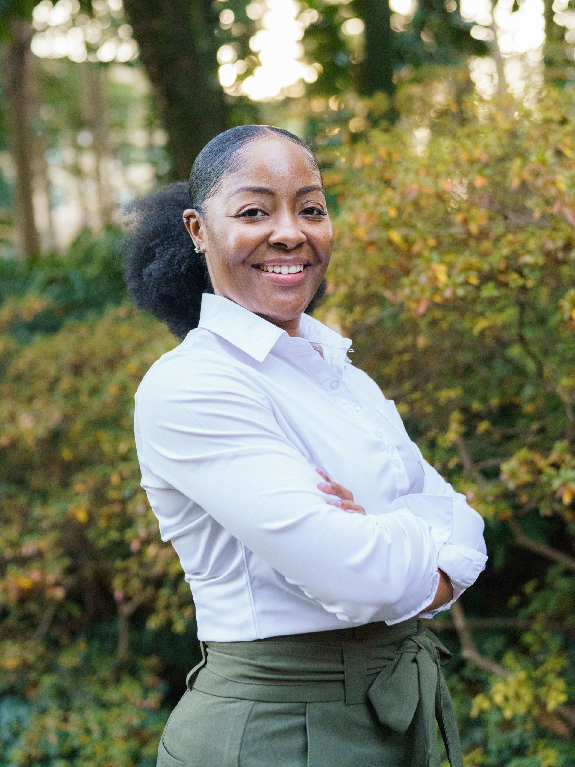 A person smiling with arms crossed, wearing a white button-down shirt and olive green pants, standing outdoors.