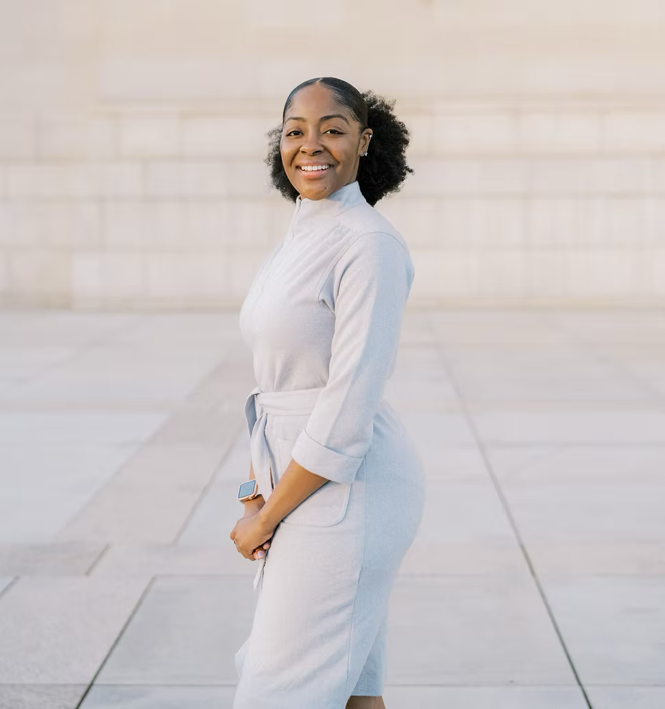 A smiling person in a light grey jumpsuit stands outdoors in front of a stone wall, posing with hands held together.