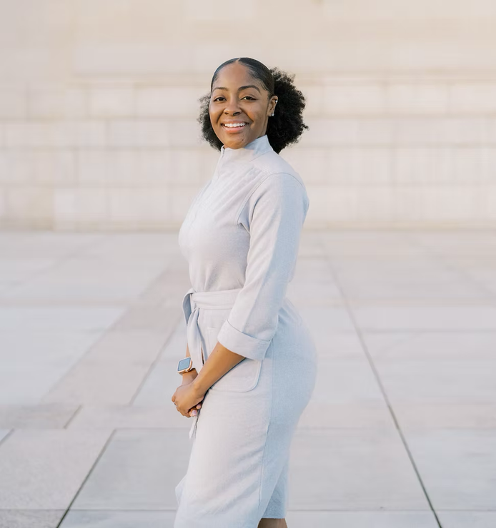 A person stands sideways, smiling at the camera, wearing a light grey belted jumpsuit against a stone-tiled background.