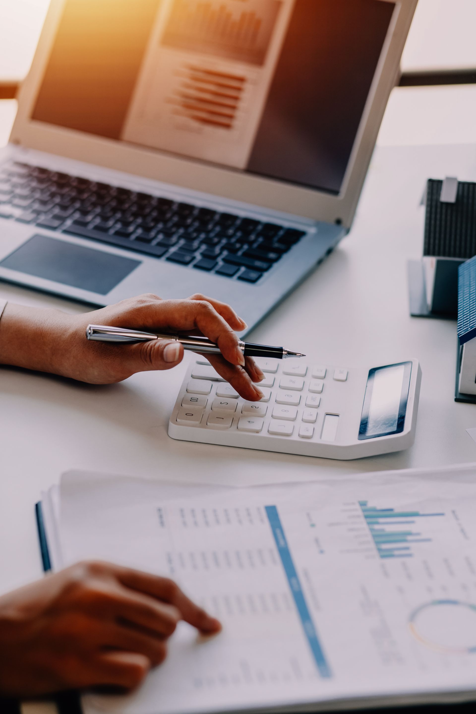 A person uses a calculator and reviews financial charts at a desk with a laptop and model houses.