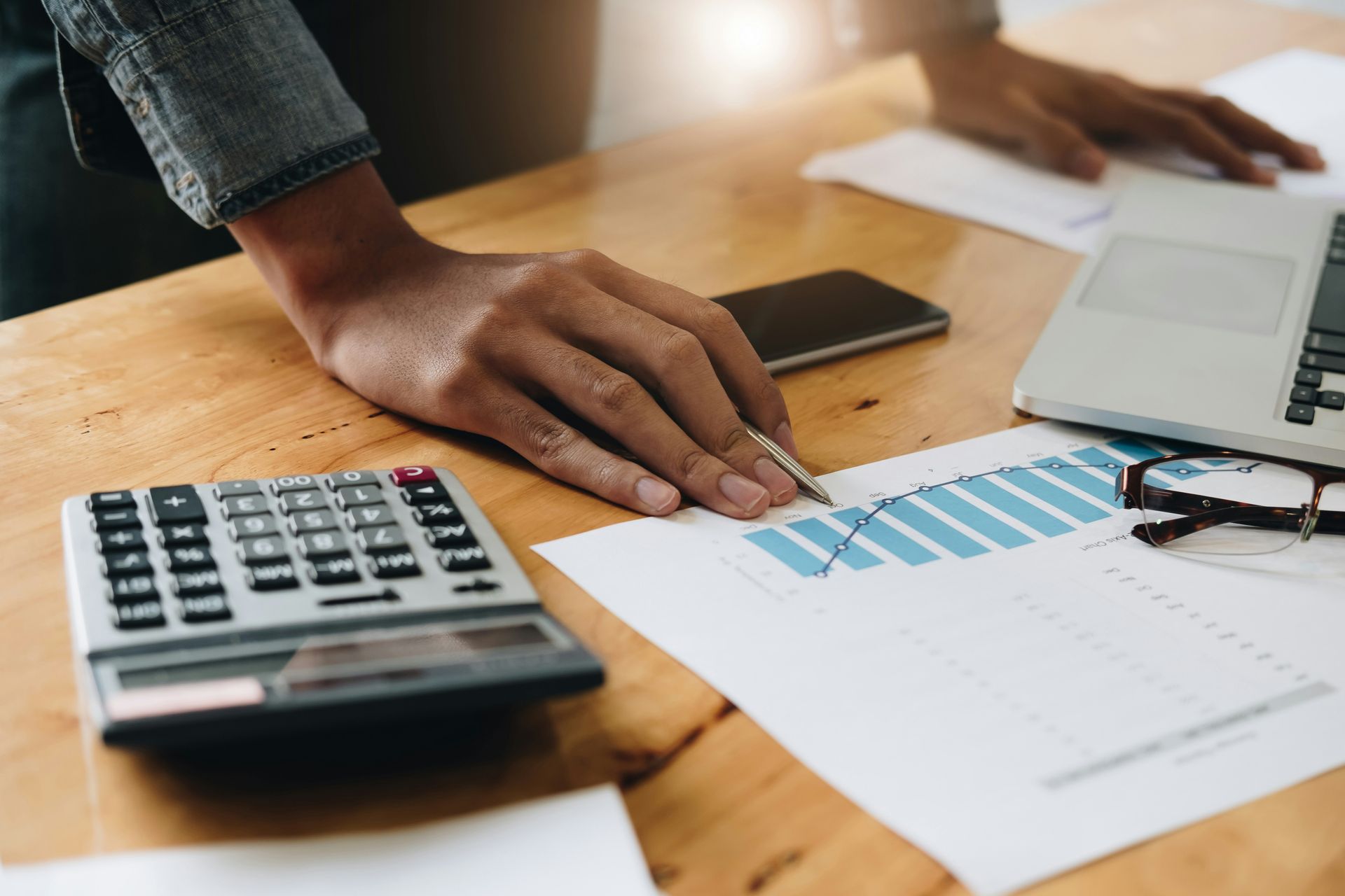 A person's hand with a pen pointing to a bar chart on a wooden desk with a calculator and laptop nearby.