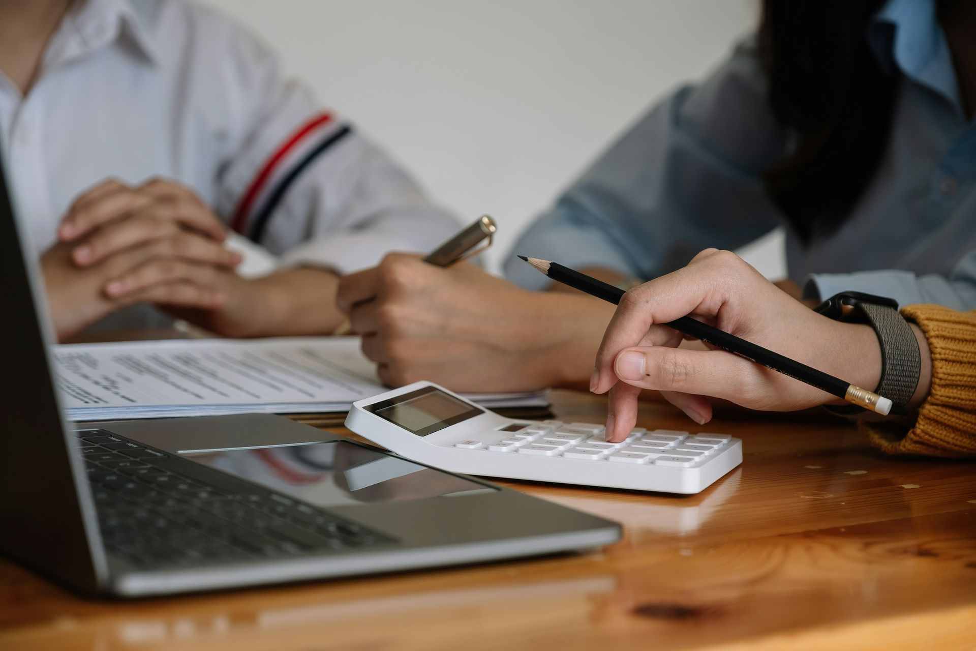 Two people work at a wooden table with a laptop, calculator, and documents while using pens to review the paperwork.
