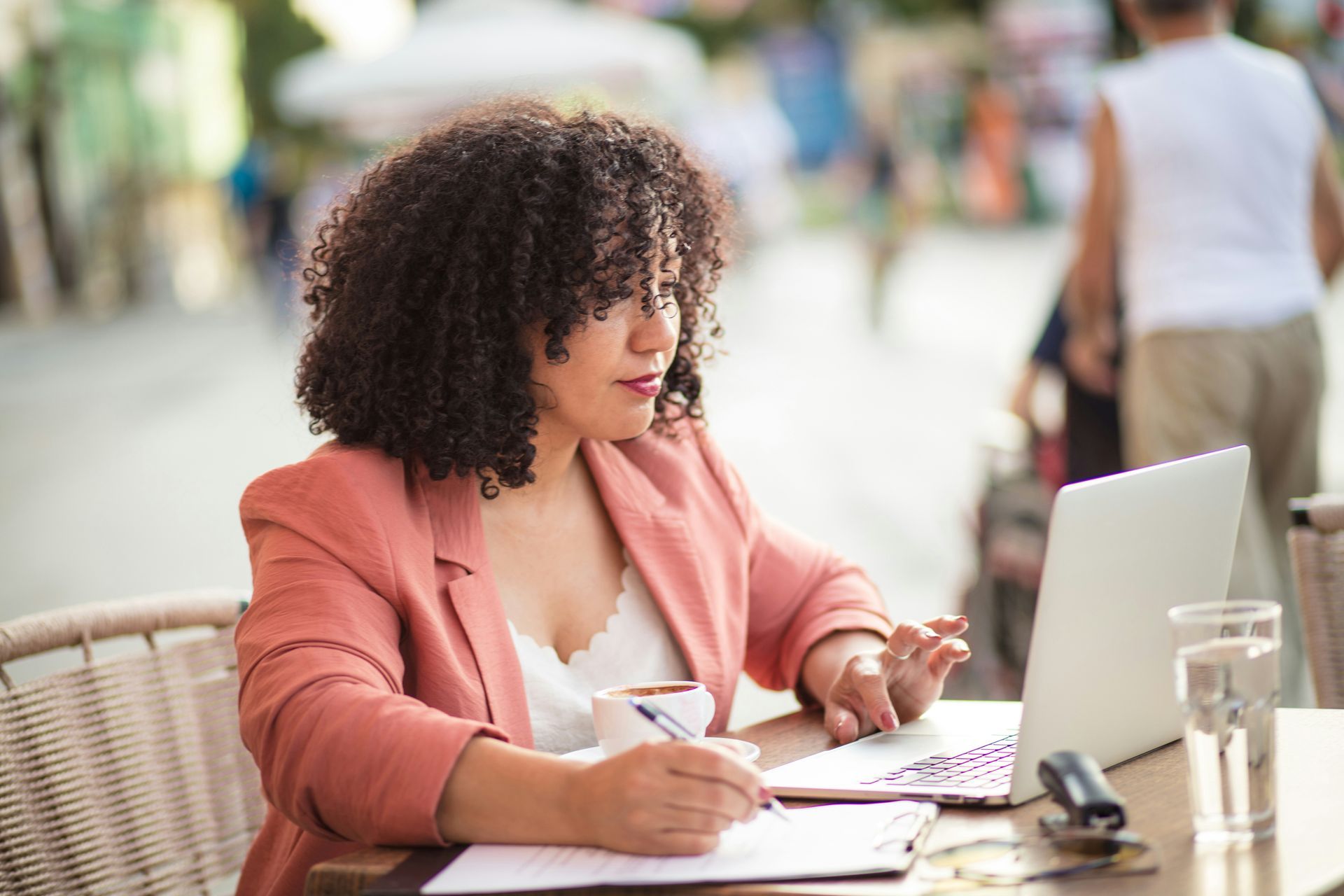 A person with curly hair wearing a pink blazer writes in a notebook while looking at an open laptop at an outdoor cafe.