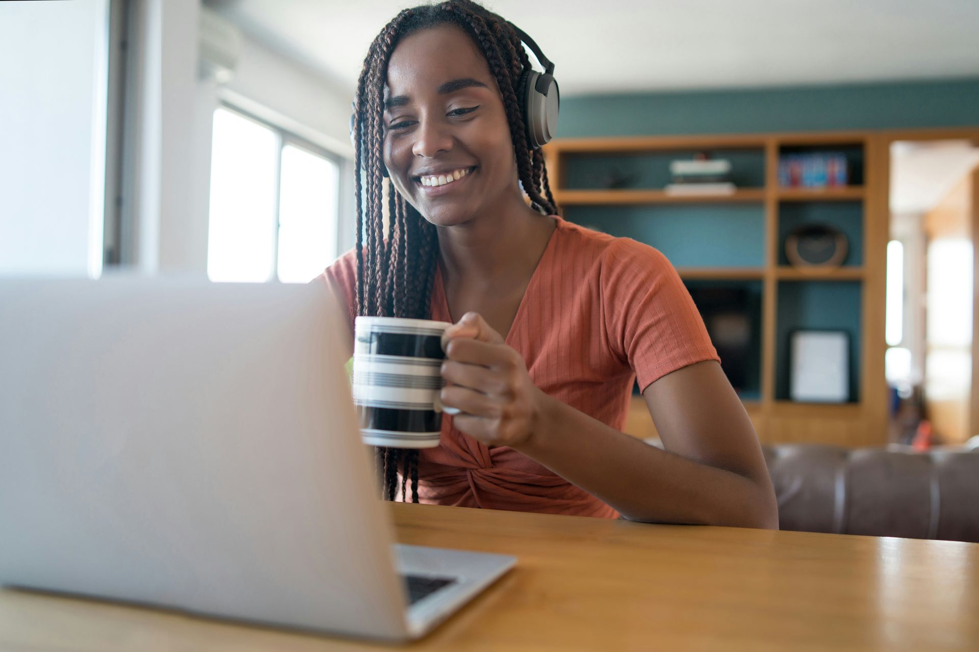 A person with long braided hair and headphones holds a striped mug while looking at a laptop in a bright living room.
