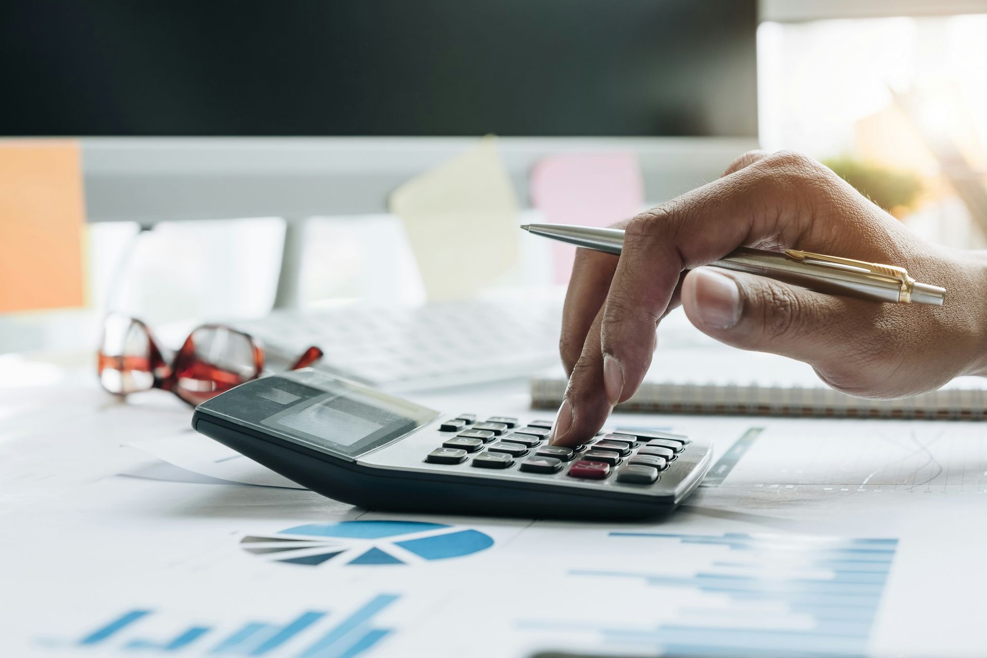 A person’s hand uses a pen to press buttons on a calculator placed over financial charts and graphs on a desk.