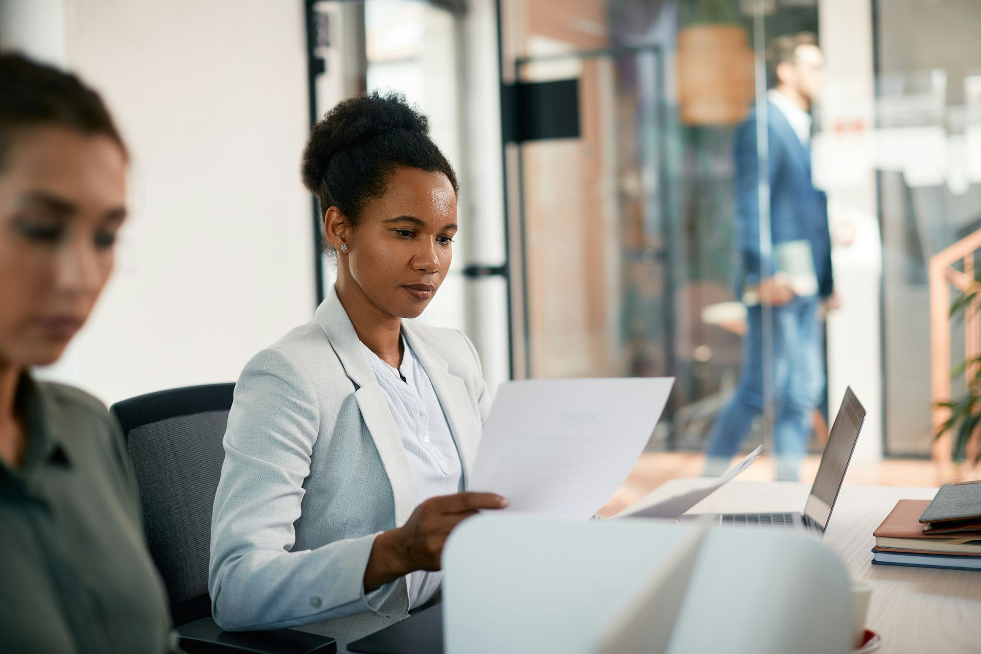 A professional at a desk reviews a document in a bright office, with another person visible in the background.