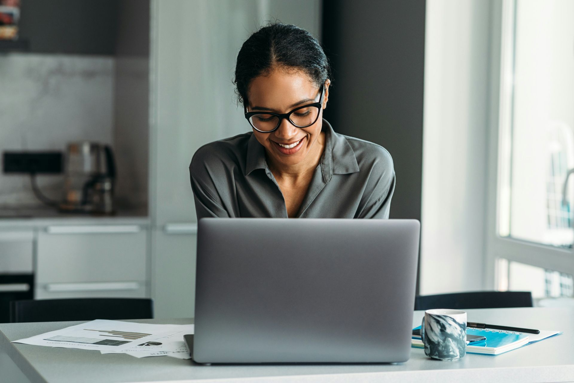 A person with glasses sits at a desk in a bright kitchen, smiling while working on an open laptop.