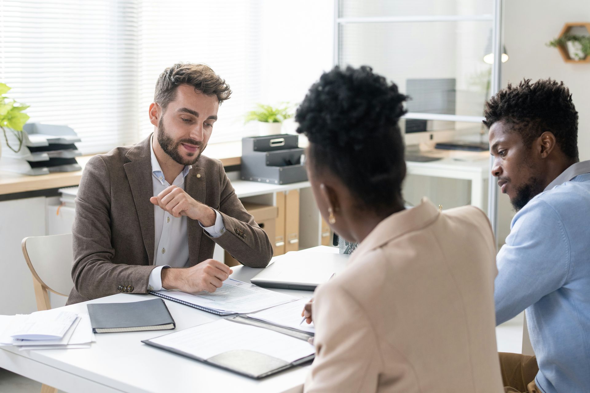 A professional advisor sits at a desk, reviewing documents with two clients in a bright office.