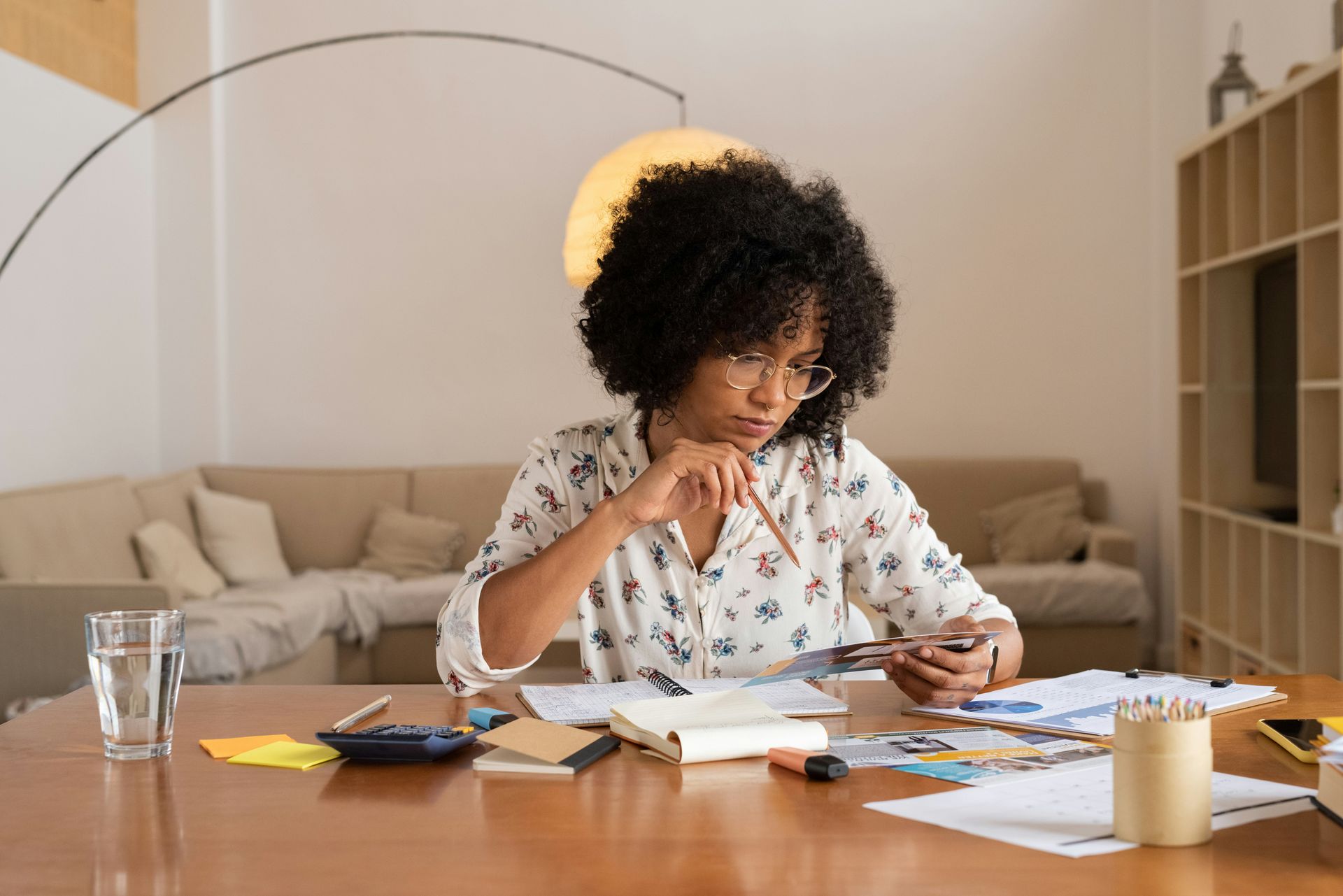 A person with curly hair sits at a wooden table, focused on reviewing papers and notes in a brightly lit living room.