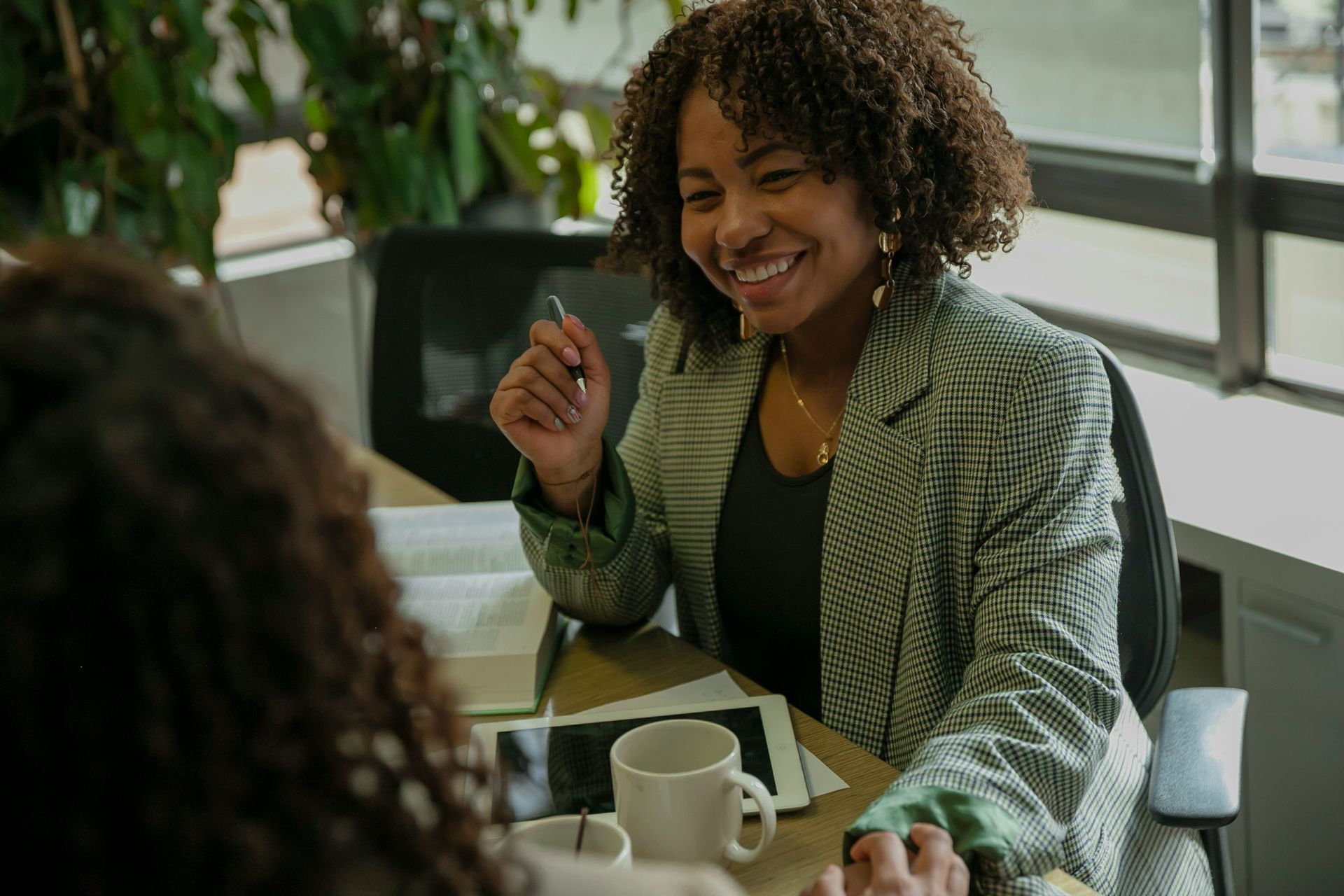 A person with curly hair smiles while sitting at a desk, holding a pen during a professional conversation.