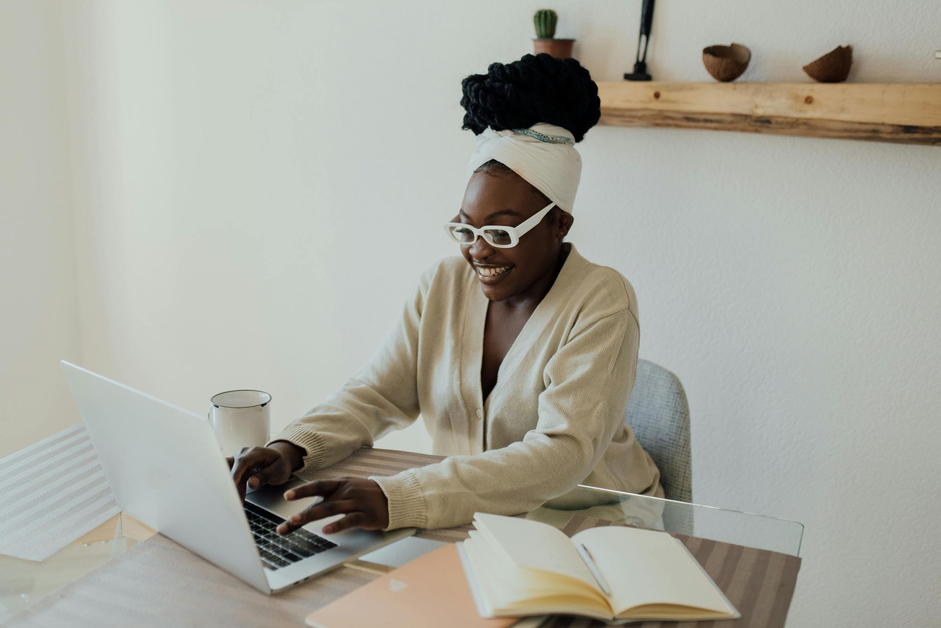 A person wearing glasses and a light-colored cardigan types on a laptop at a table with an open notebook and mug.