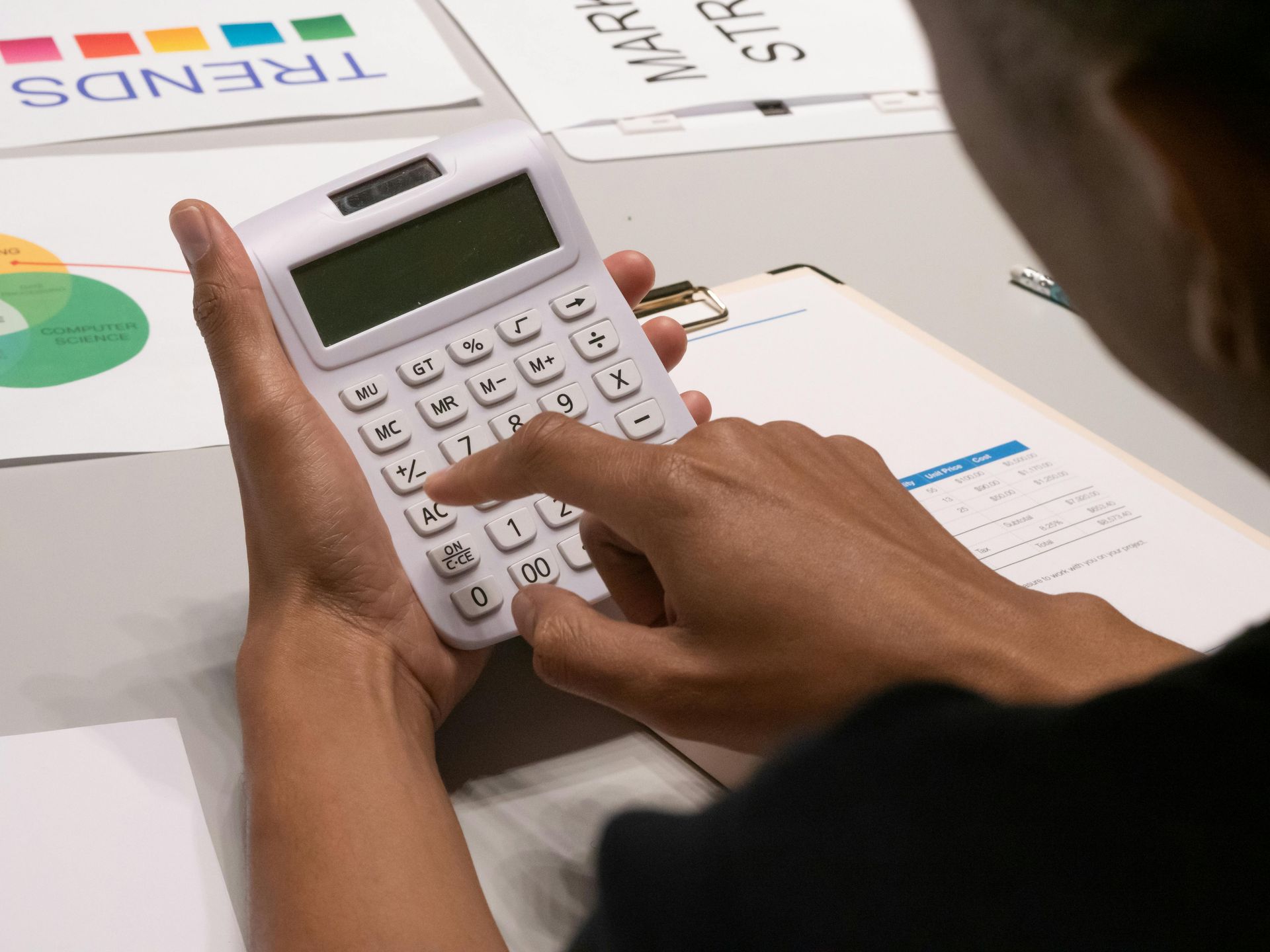 A person holds a white calculator, pressing buttons while reviewing business documents and charts on a desk.