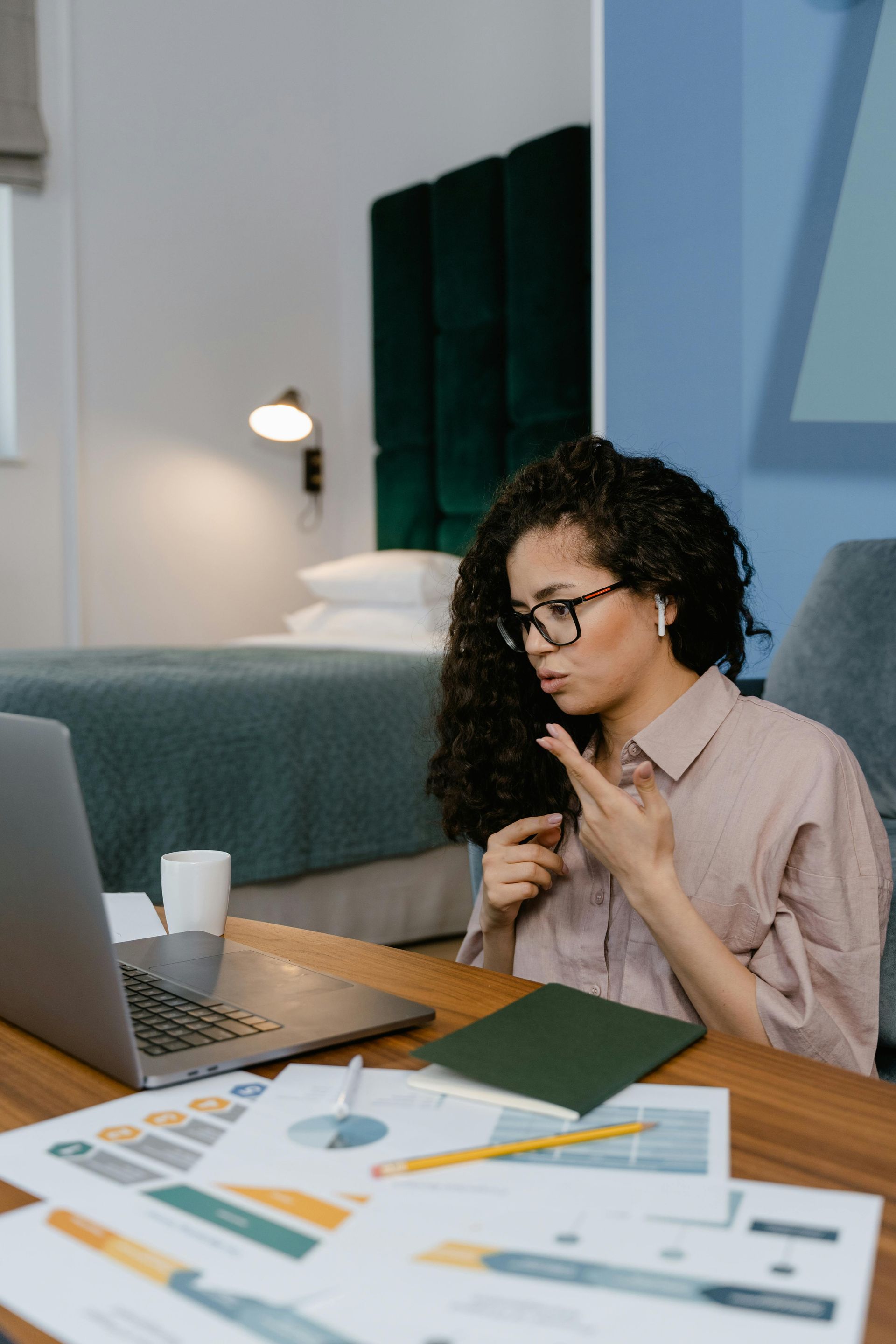 A person with curly hair and glasses sits at a desk with a laptop and charts, using hand gestures during a video call.