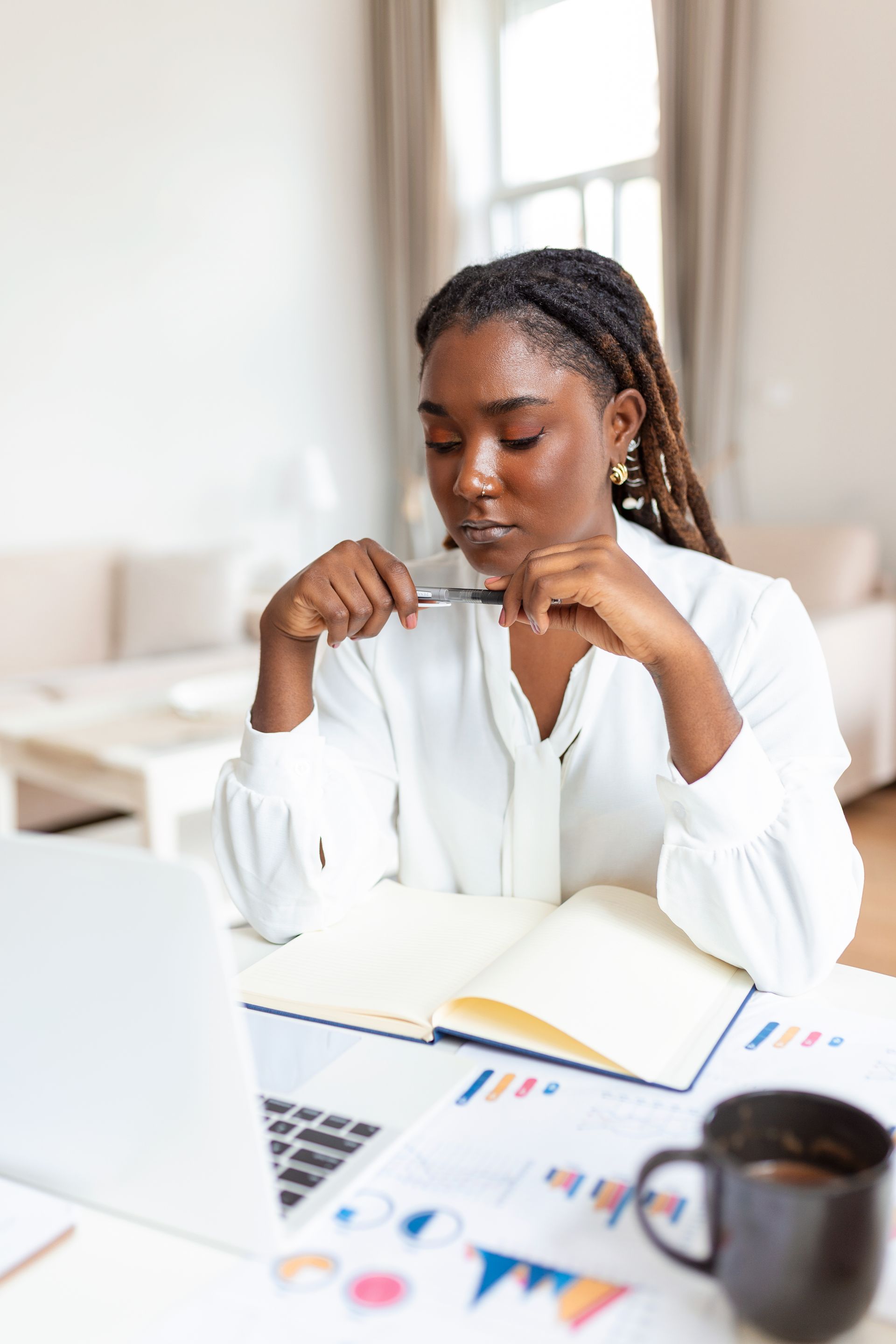 A person with braided hair sits at a desk with a laptop, open notebook, and business charts while holding a pen.