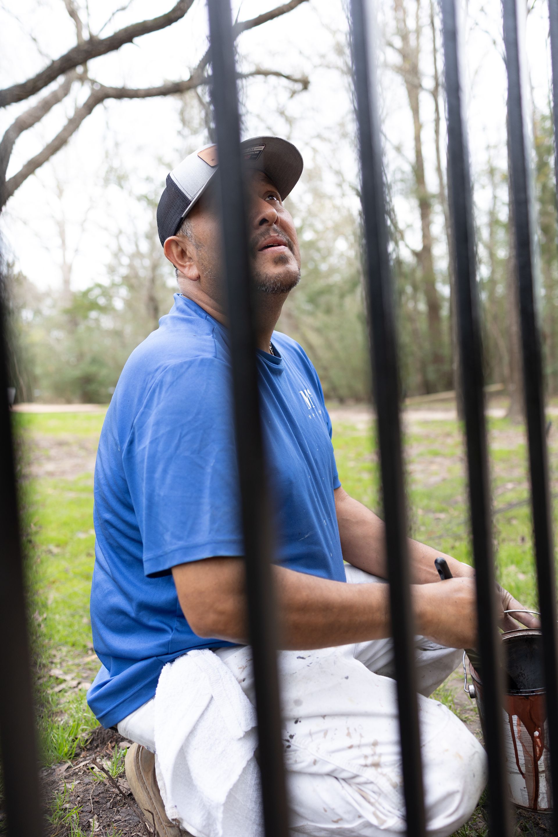 A man in a blue shirt and white pants is kneeling down in front of a fence.