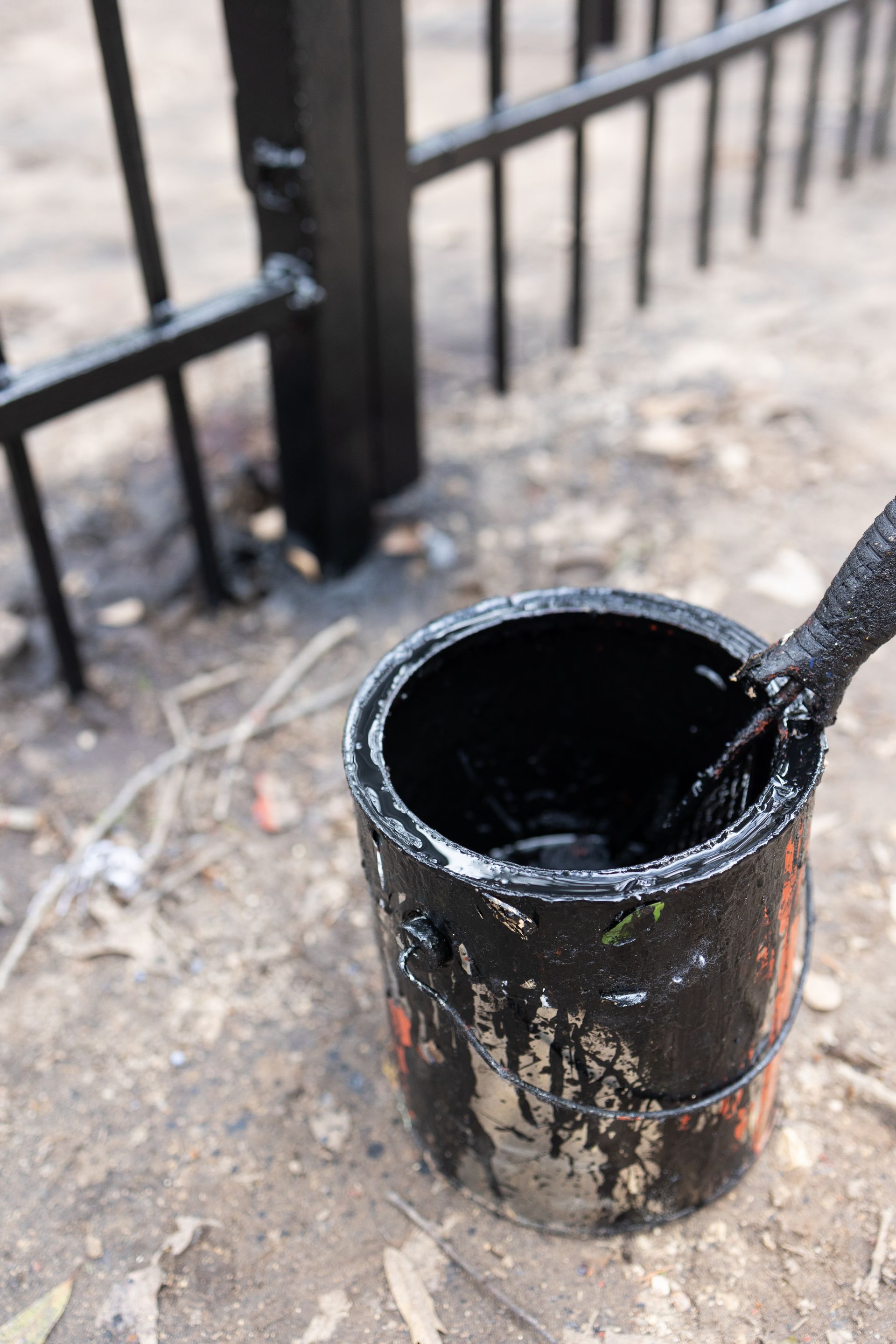 A can of black paint with a brush in it is sitting on the ground next to a fence.