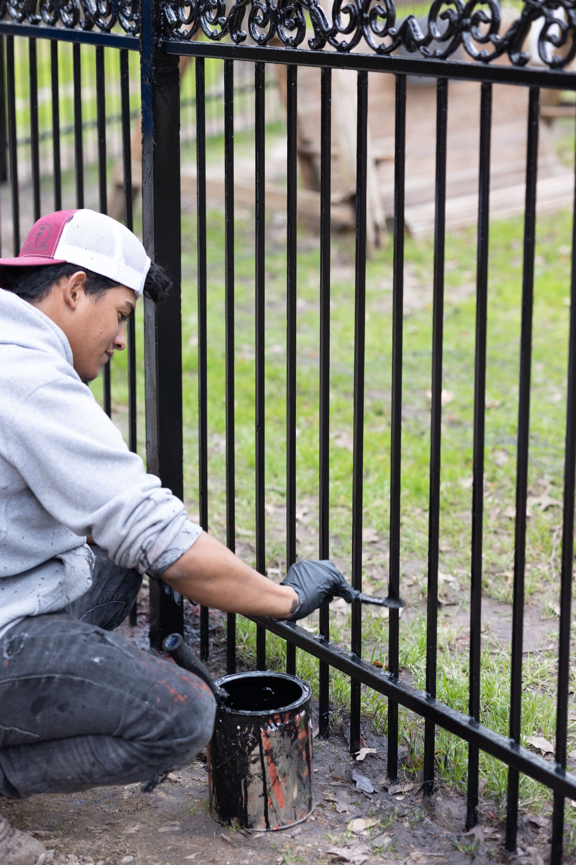 A man is kneeling down and painting a wrought iron fence.