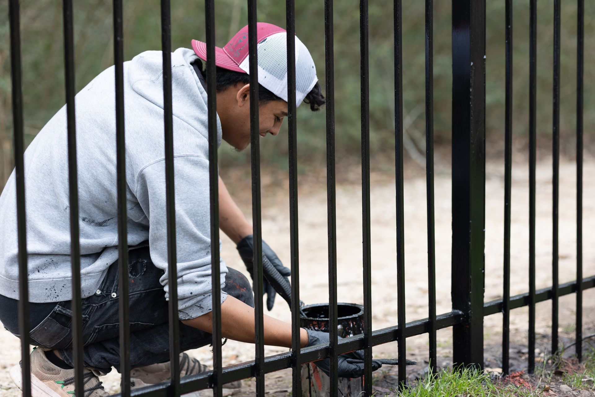 A man is kneeling down and painting a metal fence.