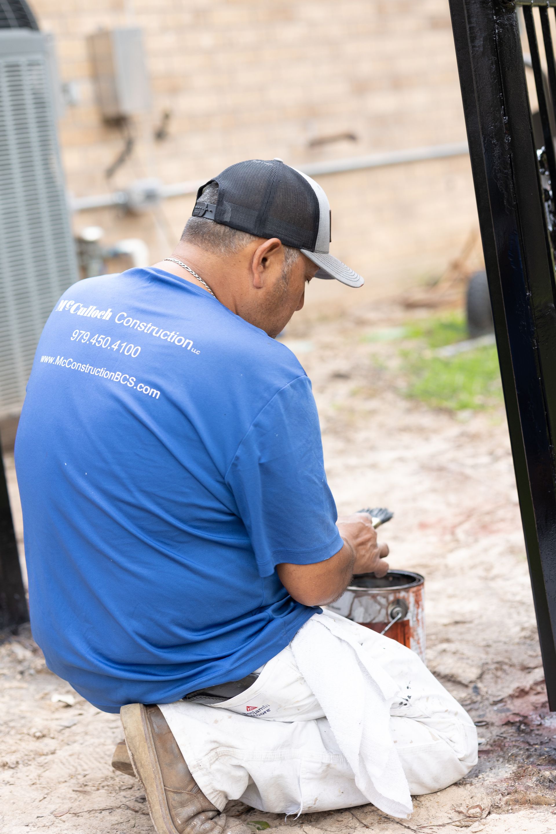 A man in a blue shirt is kneeling down and painting a fence.