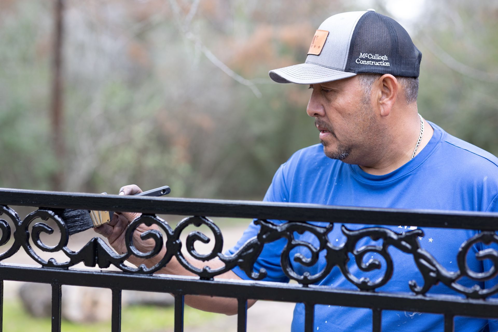 A man in a blue shirt and hat is painting a wrought iron fence.